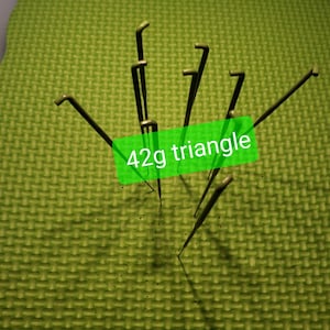 May include: A close-up of a green mat with multiple gold knitting needles arranged in a triangular shape. The text "42g triangle" is written on a green label.