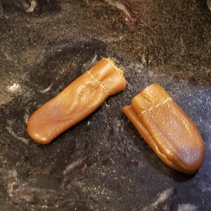 May include: Two brown leather thimbles with white stitching. The thimbles are on a dark grey speckled surface.