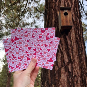 May include: A stack of cards with a pink and red heart pattern. The cards are being held in front of a tree with a birdhouse.