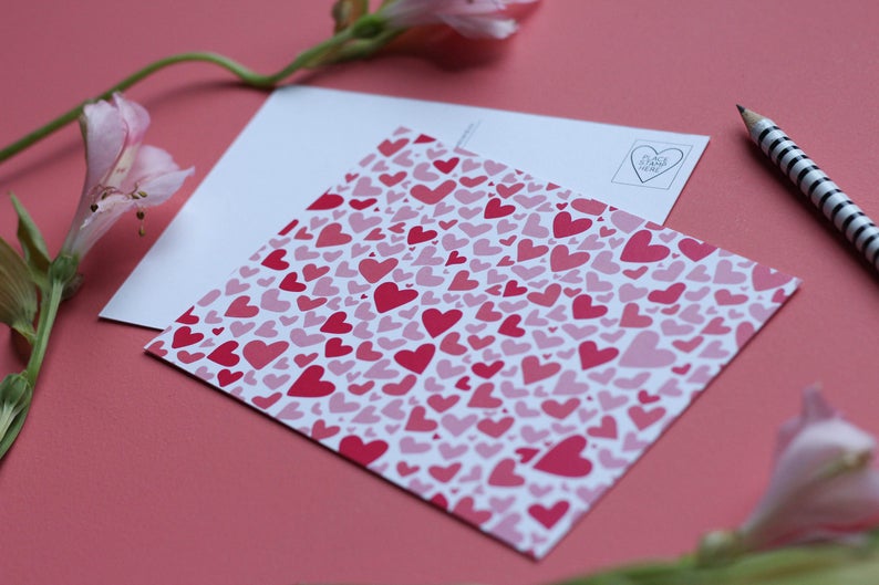 May include: A white postcard with a pink and red heart pattern. The postcard has a blank space for writing a message and a stamp with a heart and the text "I love you".