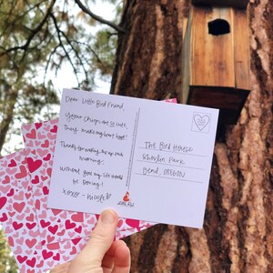 May include: A pink and white postcard with a handwritten message to a bird. The message reads "Dear Little Bird Friend, your chirps are so cute, They make my heart hoot! Thanks for waking me up each morning. Without you my life would be boring! xoxo - nicole". The postcard is sitting on a stack of pink and white cards with hearts printed on them. The postcard is in front of a tree with a birdhouse.