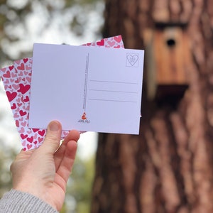 May include: A stack of pink and white postcards with a heart pattern and the text "Place Stamp Here" on the back. The postcards are held in a hand and the background is a blurry image of a tree.