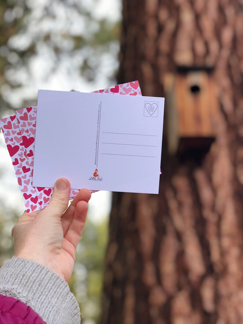 May include: A stack of pink and white postcards with a heart pattern and the text "Place Stamp Here" on the back. The postcards are held in a hand and the background is a blurry image of a tree.