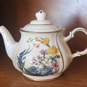 Könnte beinhalten: Eine weiße Teekanne aus Keramik mit Blumenmuster. Die Teekanne hat einen goldenen Rand um den Deckel und den Ausguss. Die Blumen sind gelb, rosa, blau und grün.