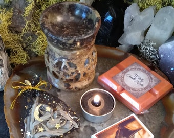 May include: A brown stone wax melt burner with a pentacle cutout, a black tea light candle, an orange wax melt bar labeled Witchy Candle Melts, a triangular black wax piece with gold stars and moon, and a matchbox with a plague doctor illustration.