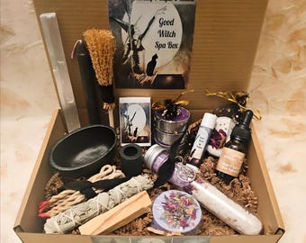 May include: A cardboard box filled with spa and spiritual items. Includes a black cauldron, sage bundles, a clear crystal, candles, a brush, a bottle of oil, a bath bomb, and a tube of bath salts. The box has a label that reads "Good Witch Spa Box."