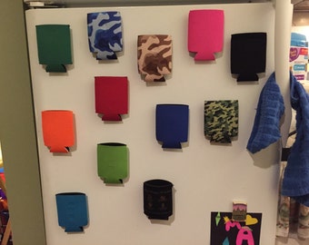 May include: A white refrigerator door with a variety of colorful can coolers attached with magnets. The coolers are in different patterns, including camouflage, stripes, and solid colors. There is a blue towel hanging on the right side of the door.