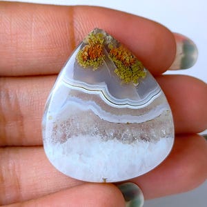 Pode incluir: Uma pedra preciosa em forma de gota com um padrão em camadas de tons brancos, cinzentos e castanhos. A parte superior da pedra apresenta manchas de laranja e verde. A superfície polida da pedra é lisa e refletora.