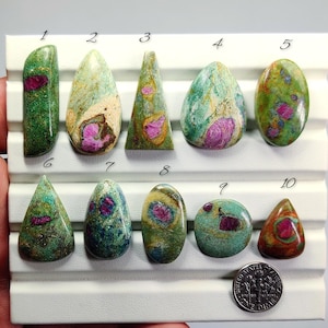 Puede incluir: Una colección de diez piedras preciosas pulidas de diversas formas, incluyendo óvalos, lágrimas y triángulos. Las piedras presentan una mezcla de tonos verdes, amarillos y rojos, con una moneda para comparar el tamaño. Las piedras preciosas se exhiben sobre una superficie blanca.