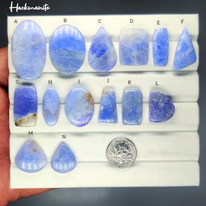 May include: A collection of blue hackmanite gemstones in various shapes, including oval, teardrop, and rectangular. The gemstones are displayed on a white surface, with a dime for size comparison. The word "Hackmanite" is at the top.