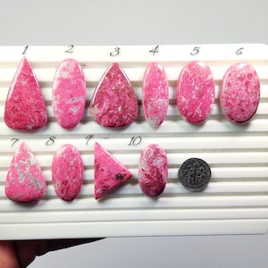 May include: A collection of ten polished rhodonite gemstones in various shapes, including oval, triangular, and teardrop. The stones display a vibrant pink hue with gray veining. A US dime is included for size reference.