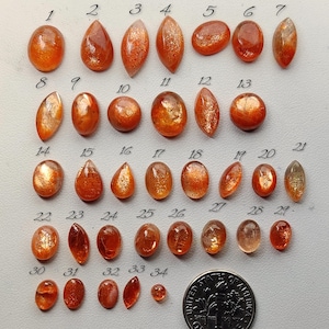 Puede incluir: Una colección de treinta y cuatro cabujones de piedra solar pulidos en varias formas, incluyendo ovaladas, de pera y marquesa, dispuestos en filas y numerados del uno al treinta y cuatro, junto a una moneda de diez centavos para escala.
