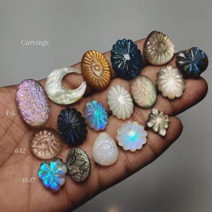 May include: A collection of small, intricately carved gemstones in various shapes including moons, flowers, and eye motifs. The stones feature iridescent, metallic, and translucent finishes in colours like blue, gold, white, and purple, labelled with text.