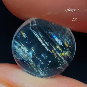 May include: A circular, translucent gemstone with a dark blue base color, featuring internal inclusions that create iridescent, multi-colored flashes of gold, blue, and silver light. The stone has a smooth, polished surface and is labeled with the number 12.
