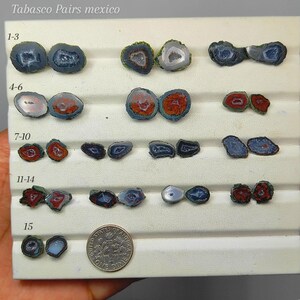 May include: A collection of Tabasco geode pairs from Mexico arranged in rows on a white display tray. The stones feature varied colors including blue, red, and grey with crystalline centers. A US dime is placed next to the final pair for scale.