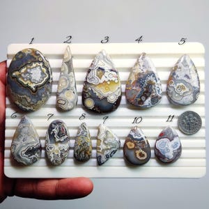 Pode incluir: Onze pedras de ágata polidas em várias formas e tamanhos, exibidas em uma bandeja branca. As pedras apresentam uma gama de cores, incluindo cinza, marrom, branco e amarelo, com padrões e texturas exclusivos. Uma moeda de dez centavos americana está incluída para referência de tamanho.