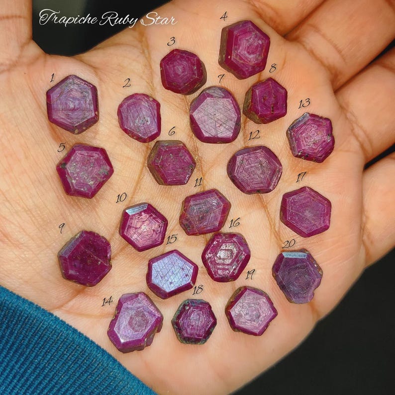 May include: A collection of twenty small, hexagonal, deep pink Trapiche Ruby Star gemstones, each labeled with a black number from 1 to 20. The stones exhibit a distinct six-rayed star pattern radiating from the center of each individual ruby.