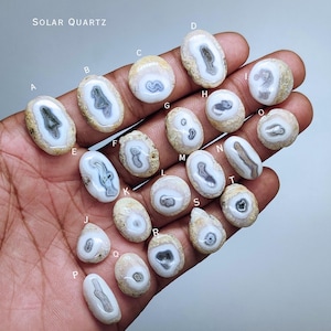 Op de afbeelding: Een verzameling zonnekwarts stenen in verschillende vormen en maten, elk met unieke patronen van grijs en wit binnen een beige rand. De tekst "SOLAR QUARTZ" is bovenaan zichtbaar.