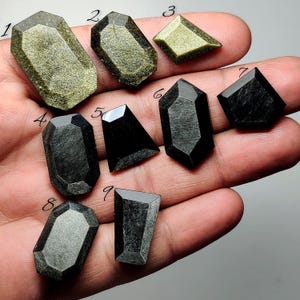Può includere: Una collezione di nove pietre preziose lucide di varie forme, tenute in una mano. Le pietre mostrano una gamma di colori, tra cui oro, nero e grigio. Ogni pietra ha un design sfaccettato unico, che mette in risalto la bellezza naturale dei minerali.