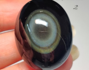 Puede incluir: Una piedra de obsidiana pulida, de forma ovalada, con un llamativo patrón similar a un ojo. La piedra presenta círculos concéntricos de tonos grises, azules y amarillos sobre un fondo negro. El texto "Swipe to see more 2" es visible.