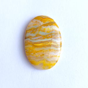 May include: Polished, oval gemstone with a layered pattern of yellow, gold, and cream hues. The stone's surface is smooth and reflective. The gemstone is displayed on a white background, accentuating its natural colours and intricate design.