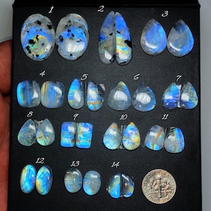 May include: A collection of fourteen polished moonstone gemstones, each with unique blue and iridescent flashes. The stones vary in shape, including oval, teardrop, and rectangular cuts. A US dime is included for size reference.