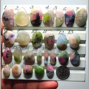 May include: A selection of twenty-six polished gemstones in diverse shapes and hues, including pink, green, and blue. The gemstones are displayed on a white tray, with a US dime for scale. The gemstones are numbered 1 to 26.