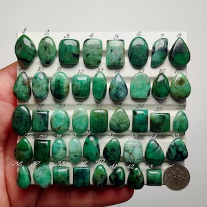May include: A collection of forty-three polished green emerald gemstones arranged in rows on a white display board. Each stone is numbered from 1 to 43, featuring various shapes including oval, square, and teardrop. A US dime is placed at the bottom right.