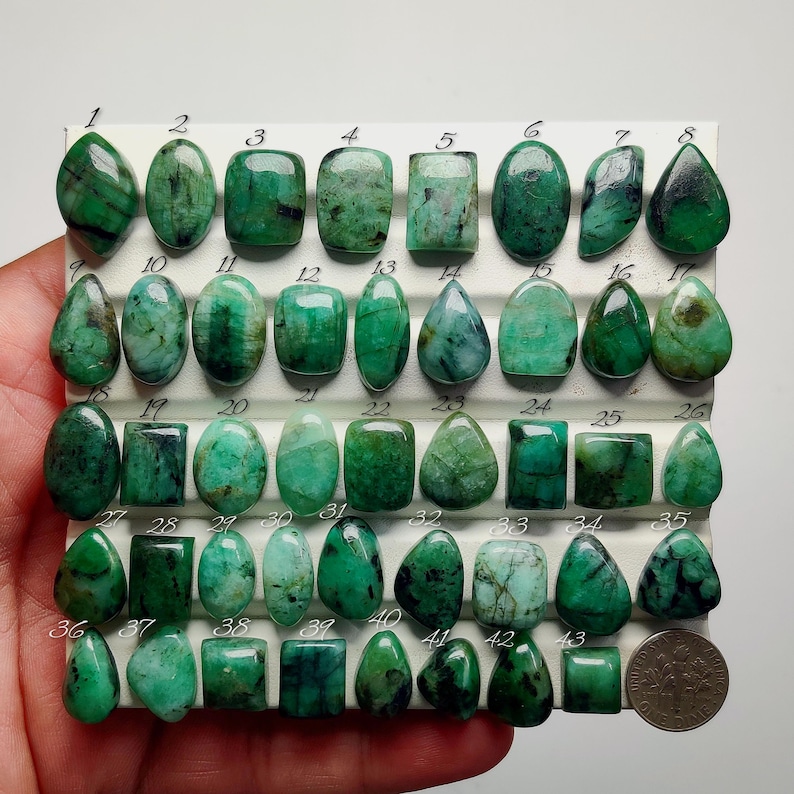 May include: A collection of forty-three polished green emerald gemstones arranged in rows on a white display board. Each stone is numbered from 1 to 43, featuring various shapes including oval, square, and teardrop. A US dime is placed at the bottom right.