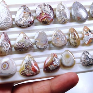 May include: A collection of polished agate gemstones in various shapes, including teardrop and oval. The stones display intricate patterns of brown, white, red, and gray, with a smooth, glossy finish. The gemstones are arranged on a white tray.