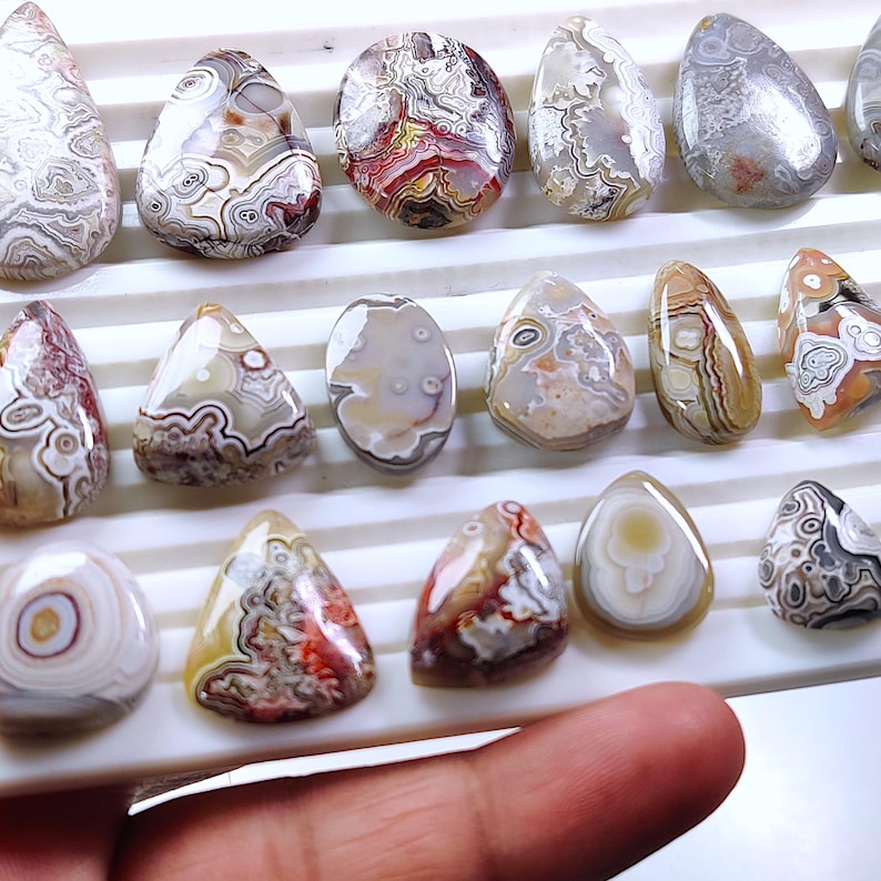 May include: A collection of polished agate gemstones in various shapes, including teardrop and oval. The stones display intricate patterns of brown, white, red, and gray, with a smooth, glossy finish. The gemstones are arranged on a white tray.