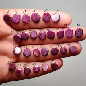 May include: A collection of twenty-seven faceted, hexagonal, deep pinkish-red rubies arranged in rows. Each gemstone is individually numbered from 1 to 27 with black text placed directly above each stone, showcasing varied natural textures.