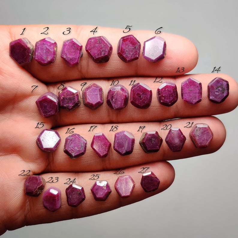 May include: A collection of twenty-seven faceted, hexagonal, deep pinkish-red rubies arranged in rows. Each gemstone is individually numbered from 1 to 27 with black text placed directly above each stone, showcasing varied natural textures.