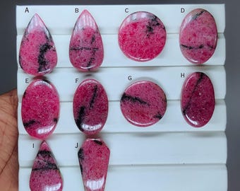 May include: A collection of bi-color rhodonite gemstones in various shapes, including teardrop, oval, and round. The stones display a vibrant pink hue with black veining. The text "BI COLOUR RHODONITE" is at the top.