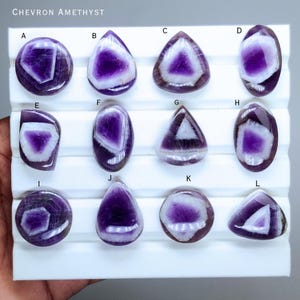 Può includere: Gemme di ametista Chevron in varie forme, tra cui rotonde, a goccia e triangolari. Le gemme presentano un sorprendente motivo di colore viola e bianco. L'immagine mostra dodici pietre individuali, ognuna con venature interne uniche.