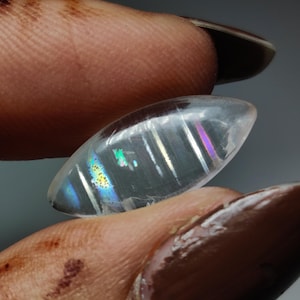 May include: A clear, oval-shaped gemstone with iridescent rainbow flashes. The stone is held between fingers, showcasing its polished surface and internal light play. The gem's colors include blues, greens, and purples, creating a captivating display.