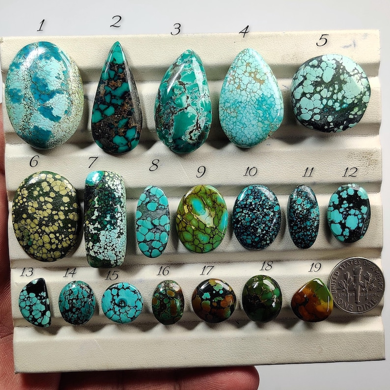 Op de afbeelding: Een verzameling van negentien gepolijste turkoois cabochons in diverse vormen en tinten blauw en groen, gerangschikt op een displaytray met genummerde labels van 1 tot 19, vergezeld door een Amerikaanse dime voor de schaalvergroting.