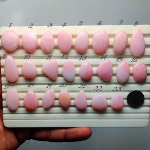 May include: A collection of twenty-three smooth, polished pink cabochon gemstones arranged on a white grooved display tray. Each stone is numbered from 1 to 23, featuring various oval, teardrop, and rounded shapes in shades of soft pastel pink.