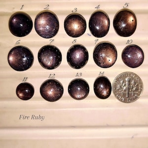 Peut inclure: Quatorze pierres précieuses cabochons rondes et ovales, brun foncé et noires, numérotées de un à quatorze et disposées en rangées. Une pièce de dix cents américaine est placée à côté de la quatorzième pierre. Le texte Fire Ruby est imprimé.