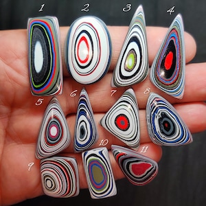 May include: A collection of eleven polished stones with colorful, concentric patterns. The stones vary in shape, including oval, teardrop, and rectangular. The designs feature a mix of colors like red, blue, black, and white, creating a striking visual effect. These stones could be used for jewelry or decorative purposes.