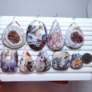 Puede incluir: Una colección de nueve piedras preciosas de ágata pulidas en varias formas, incluyendo lágrimas, óvalos y rectángulos. Las piedras muestran una gama de colores, como marrón, blanco, rojo y púrpura, con patrones intrincados. Se incluye una moneda como referencia de tamaño.