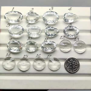 Puede incluir: Una colección de gemas claras y facetadas en varias formas, incluyendo cortes ovalados, de pera y redondos. Se incluye una moneda para la comparación de tamaños. Las gemas se muestran sobre una superficie blanca.