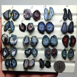 May include: A collection of nineteen pairs of small, polished agate slices arranged on a white display board, each numbered from 1 to 19. The stones feature varied colors including blue, red, and black with crystalline centers. A US dime is included for scale.