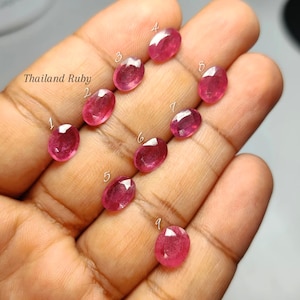 May include: Nine oval-cut Thailand Ruby gemstones arranged on a palm, each labelled with a white number from one to nine. The faceted stones exhibit varying shades of deep pink and red, showcasing a polished, reflective surface and clear clarity.