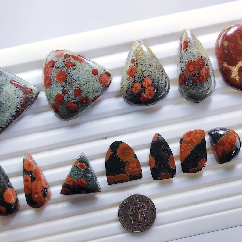 May include: A collection of polished gemstones in various shapes, including teardrop, triangular, and oval. The stones feature a mix of colors, including red, black, and gray, with unique patterns. A US dime is included for size reference.