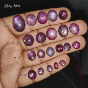Puede incluir: Una mano muestra 21 piedras preciosas de rubí estrella en varias formas ovaladas y redondas. Las gemas varían en color de rojo intenso a rosa púrpura, con un efecto de estrella visible en algunas. El texto "Ruby Star" está arriba.