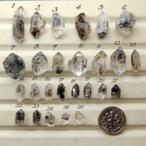 Puede incluir: Una colección de veintiséis pequeños cristales de cuarzo transparentes y ahumados dispuestos en filas sobre una bandeja de exhibición, numerados del uno al veintiséis. Una moneda de diez centavos de EE. UU. se sitúa en la esquina inferior derecha.