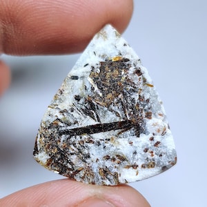 Op de afbeelding: Een driehoekige edelsteen met een witte basis, bespikkeld met bruine en zwarte minerale insluitsels. De steen wordt tussen twee vingers gehouden, waarbij de natuurlijke patronen en texturen worden getoond. De edelsteen heeft een gepolijst oppervlak.