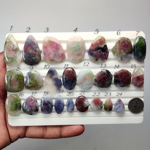 May include: A collection of twenty-four polished, multi-colored gemstones arranged on a white display tray. Each stone is numbered from 1 to 24, featuring various shades of pink, green, blue, and white. A silver coin is placed next to number 24.