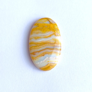 May include: An oval gemstone with a polished surface. The stone displays a layered pattern of yellow, white, and tan colours. The gemstone's design is reminiscent of natural stone formations, with a smooth, glossy finish.